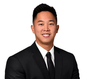 Professional headshot of a smiling Asian man in a black suit with white shirt and black tie on a transparent background