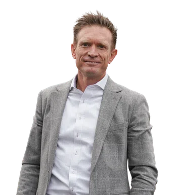 Man wearing a gray plaid blazer, white dress shirt, blue jeans, and a large decorative belt buckle smiling