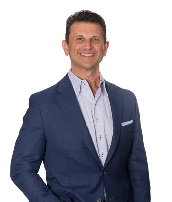 Smiling man in a blue blazer, light dress shirt, and jeans with hands in pockets against a white background