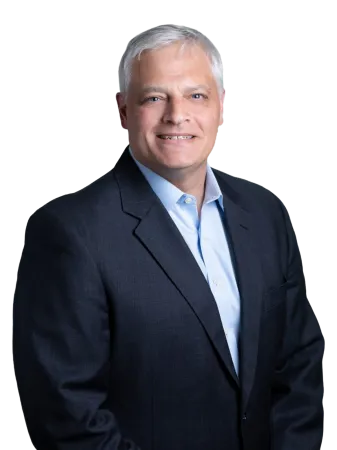 Smiling middle-aged man with gray hair wearing a dark suit jacket and light blue shirt on black background