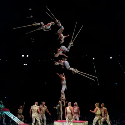 Acrobats perform a high-flying trapeze routine above performers on stage in a dark theater.