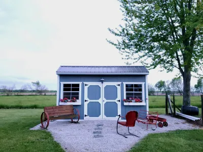 Blue garden shed with white trim, flower boxes, vintage red wagon, and outdoor seating on gravel patio.
