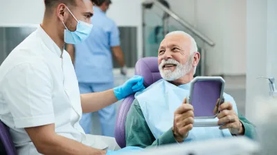 Older man smiling and holding a mirror at dental office while dentist in mask talks to him.