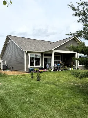 Single-story gray house with white trim, covered patio, lawn, potted plants, and outdoor seating under cloudy sky