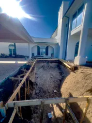 Sunlit backyard with a deep rectangular pool excavation beside a white modern house under clear blue sky.