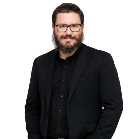 Man with glasses, beard, and black suit smiling confidently against a black background