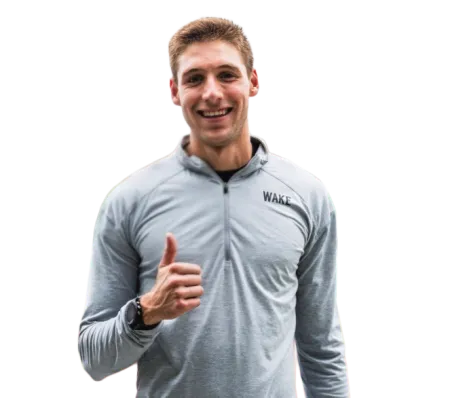 Smiling young man in gray WAKE sweatshirt giving thumbs up, standing confidently with black watch and striped pants.