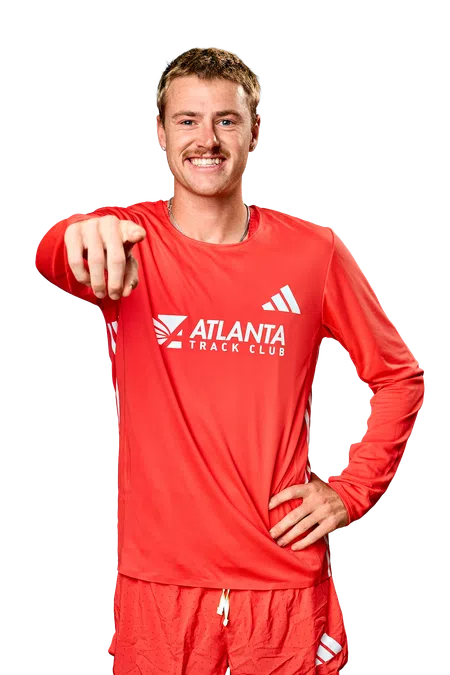 Smiling male athlete in red Atlanta Track Club outfit pointing forward with hand on hip against black background