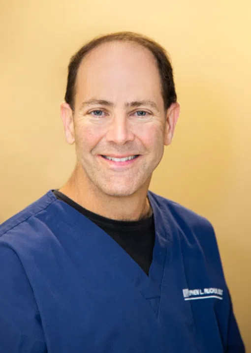Smiling middle-aged man in blue medical scrubs with light brown background, facing the camera.