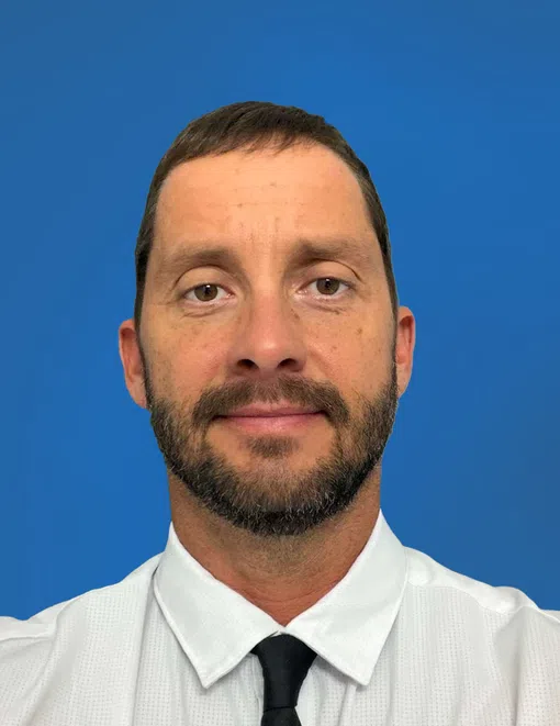 Middle-aged man with short gray hair wearing a dark suit and white shirt against a blue background.