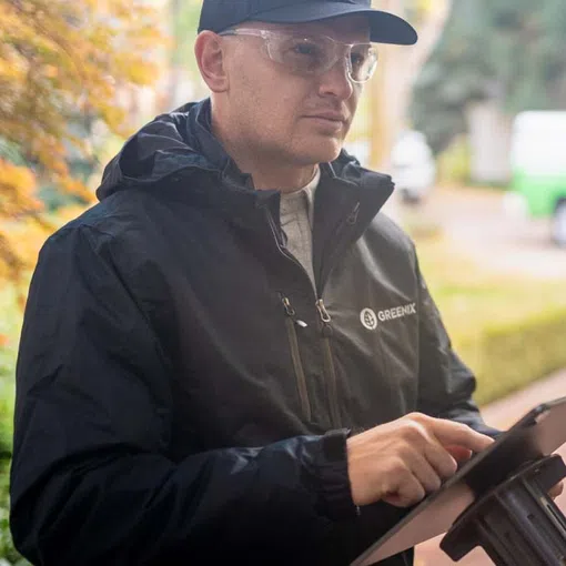 Man wearing safety glasses and a black Greenix jacket uses a tablet outdoors with autumn foliage in the background