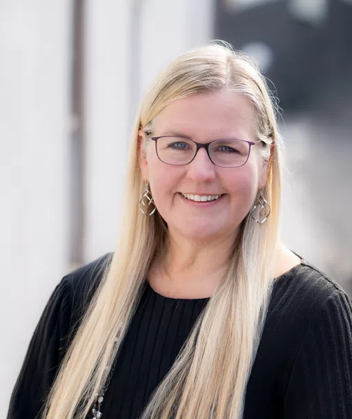 Smiling woman with long blonde hair, glasses, black top, and silver jewelry in a soft outdoor setting.