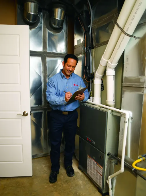 Technician inspecting HVAC system and taking notes on a clipboard in a utility room with pipes and ductwork.