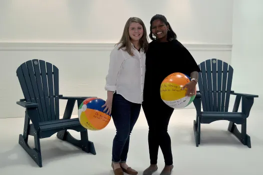 Two alums hug holding beach balls in front of blue chairs.