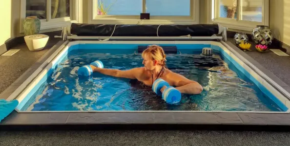 Woman exercising with aqua dumbbells in an indoor swim spa surrounded by windows and decorative items