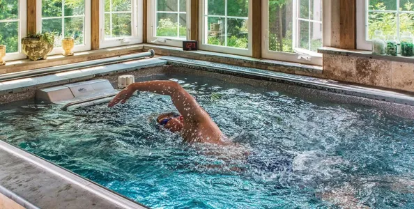 Man swimming laps in an indoor pool surrounded by large windows with natural light and plants outside.