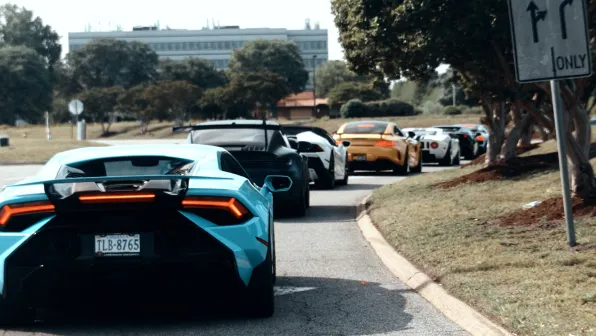 Line of high-performance sports cars driving on a curved road beside a grassy park under a clear sky