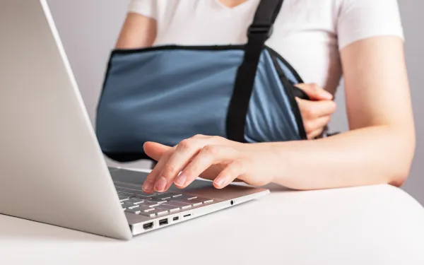 Person with arm in sling typing on a laptop at a white table, showing injury and work activity.