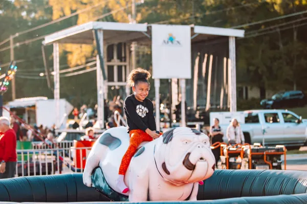 Smiling girl riding a white and black inflatable bulldog at an outdoor fair with people and vehicles in the background