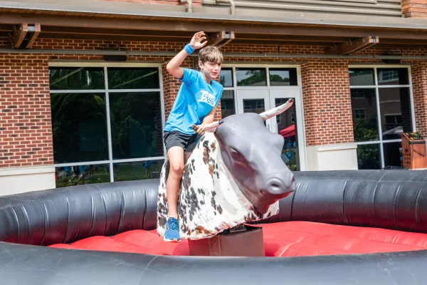 Young boy riding a mechanical bull at an outdoor event near a brick building on a sunny day.