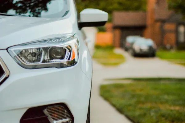 Close-up of a white car's headlight and side mirror parked on a residential street with houses in the background