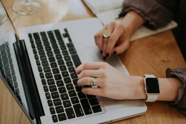 Woman typing on a laptop keyboard with rings and a smartwatch on wooden table.