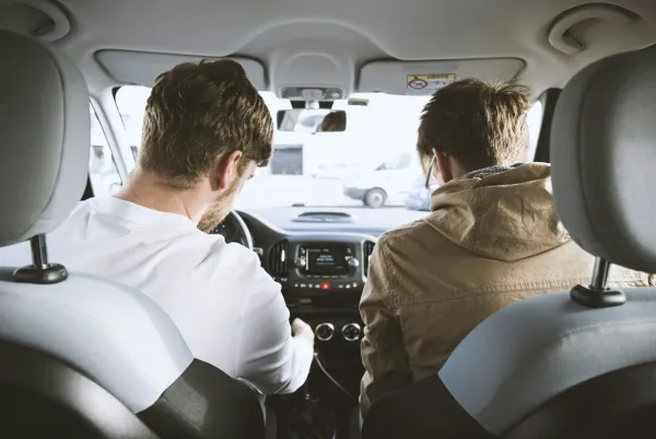 Two men sitting in a car's front seats, looking down at the dashboard and controls during daytime.