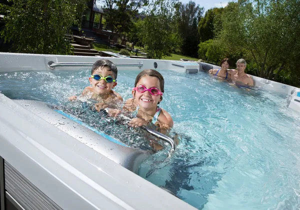 Two children wearing swim goggles swim in a backyard swim spa with adults relaxing in the background