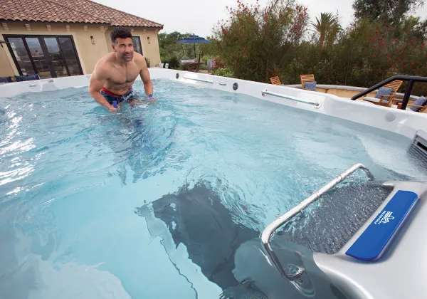 Man swimming on an underwater treadmill in an outdoor swim spa near a house with patio furniture outside.