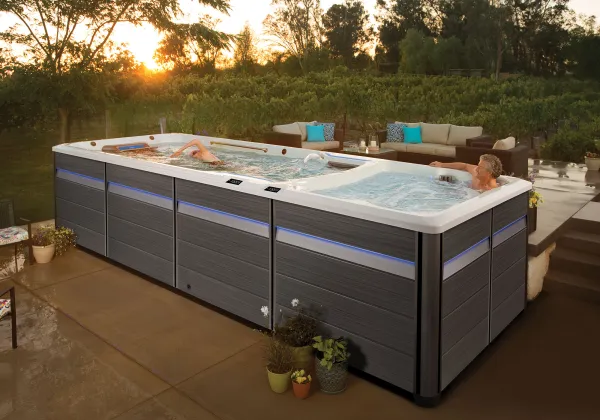 Modern outdoor swim spa with two people relaxing, surrounded by garden plants and patio furniture at sunset.
