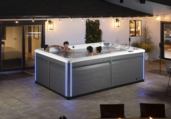 Four people relaxing in a modern hot tub with blue LED lights on a tiled patio at dusk.