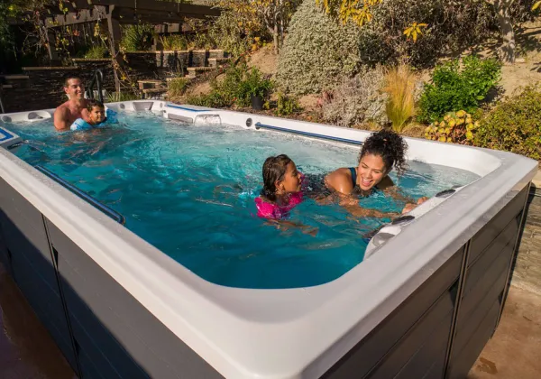 Family enjoying a swim in a large outdoor swim spa with clear blue water surrounded by greenery.