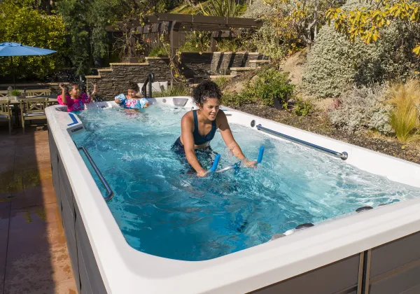 Woman exercising on an underwater bike in a swim spa with two children playing in the background outdoors.