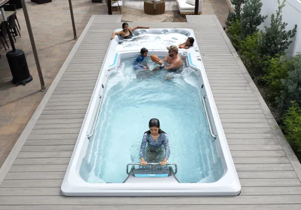 Family enjoying swim spa with woman exercising and others relaxing in jets outdoors on wood deck.