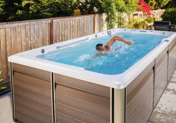 Man swimming in a modern outdoor swim spa with brown panels and wooden fence background