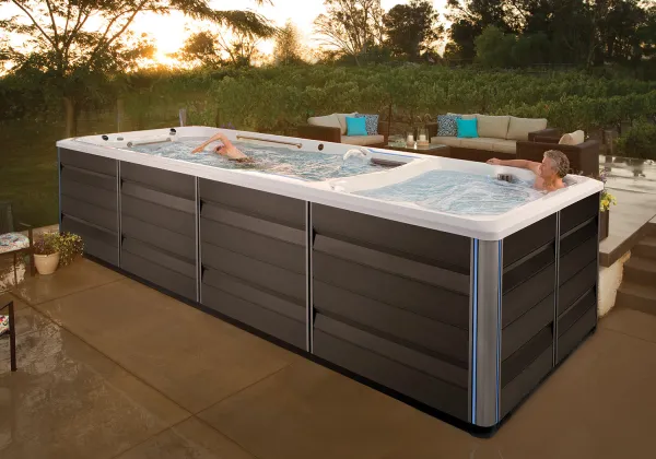 Outdoor swim spa with two people swimming and relaxing in a backyard patio setting at sunset.