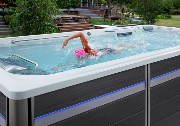 Person swimming freestyle in a modern outdoor swim spa with blue LED lights in a backyard.