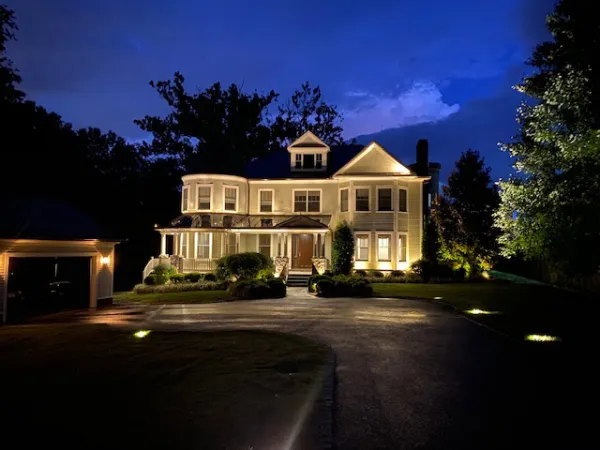 Large illuminated two-story house with wraparound porch and driveway at night with dark blue sky.