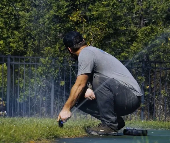 Man in gray shirt crouching while watering grass in a sunny outdoor area with trees and fence