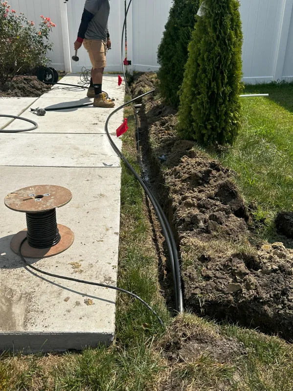 A person digging a trench for wiring installation in a residential yard with grass and trees.