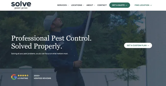 Pest control professional treating exterior with spray equipment near house surrounded by trees.