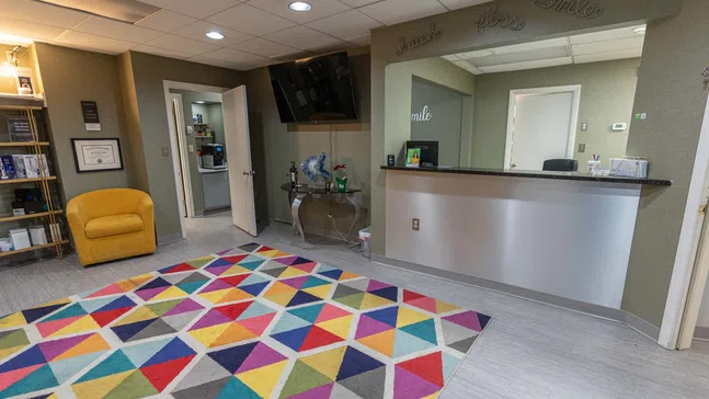 Bright dental office reception with colorful geometric rug, yellow chair, and modern desk under 'smile' decor.
