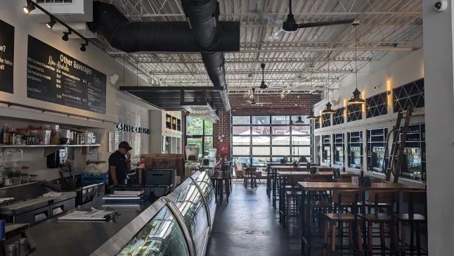 Modern café interior with high ceilings, wooden tables, glass display case, and industrial lighting.