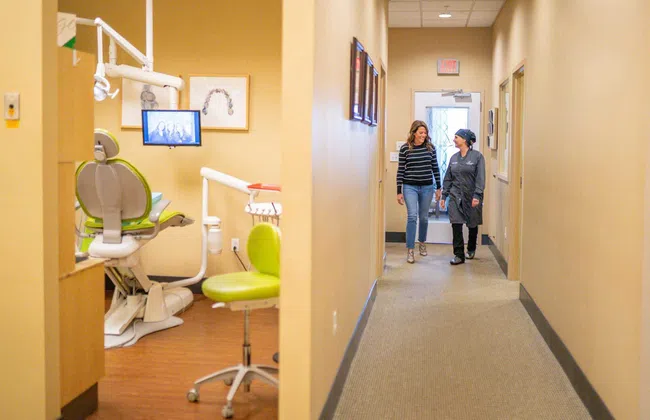 Two women walking and talking in a dental clinic hallway with a green dental chair and equipment visible in a treatment room.