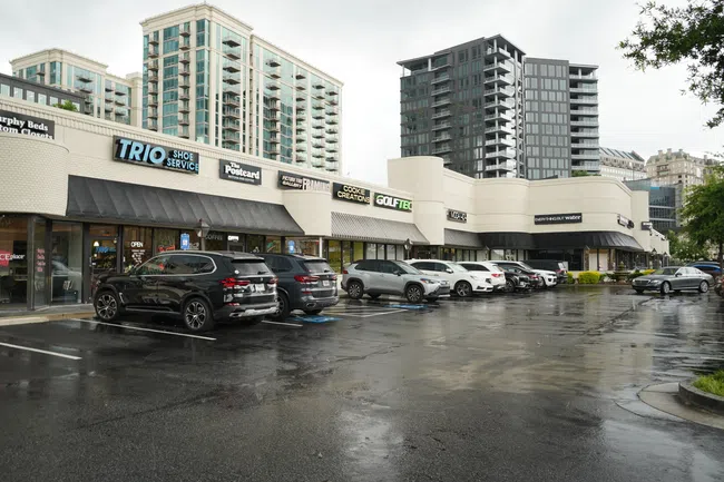 Strip mall with multiple stores and parked cars on a wet day, tall apartment buildings in the background