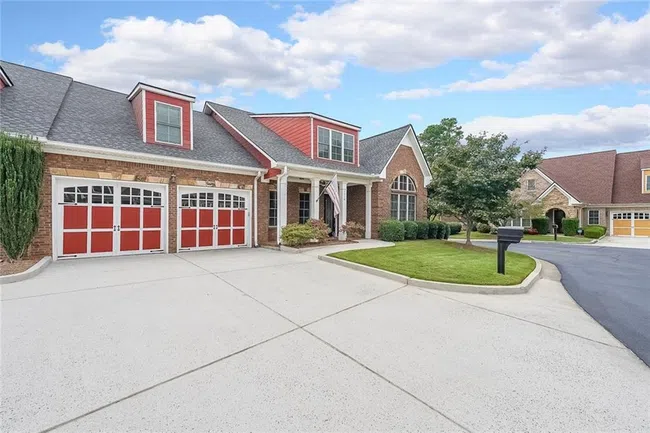 Suburban brick house with red accents, three-car garage, manicured lawn, and clear blue sky.