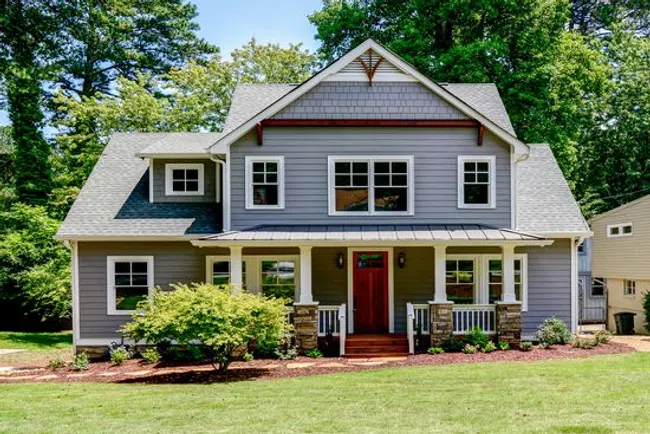 Two-story gray house with white trim, a red front door, stone pillars, and a manicured lawn in a wooded area.