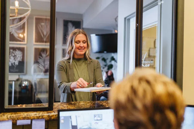 Receptionist smiling as she hands a clipboard to a patient at the front desk in a modern office.