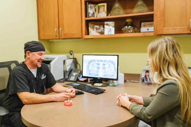 Dentist explaining dental mold to female patient at office desk with dental x-ray on computer screen.