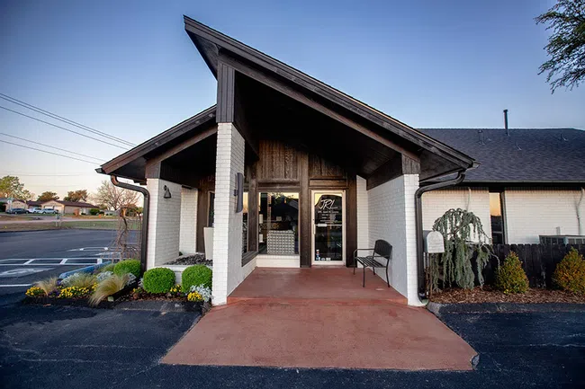 Modern building entrance with steep angled roof, white brick walls, glass door, and surrounding greenery at dusk.